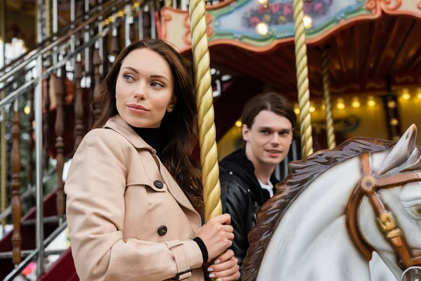 Pretty woman in trench coat riding carousel horse near blurred boyfriend in amusement park — Stock Photo