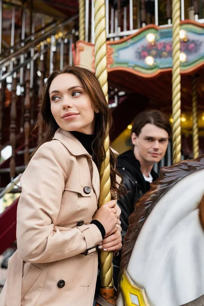 Pretty young woman in trench coat riding carousel horse near blurred boyfriend — Stock Photo