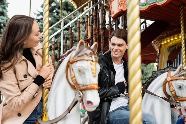 Jovem alegre olhando para namorada montando cavalo carrossel no parque de diversões — Fotografia de Stock