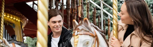 Веселый молодой человек глядя на подругу верхом на карусели лошадь в парке развлечений, баннер — стоковое фото
