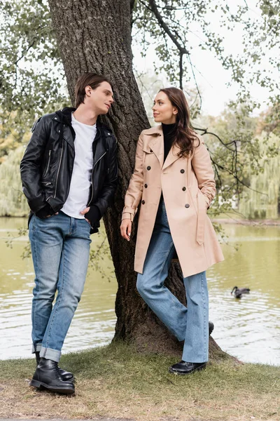 Full length of stylish man and pretty woman in trench coat looking at each other near river in park — Stock Photo