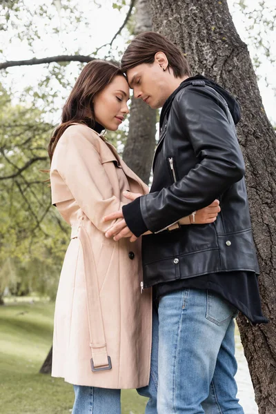 Side view of sensual young man and pretty woman in trench coat hugging near tree in park — Stock Photo