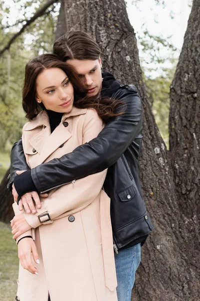 Joven en chaqueta negra abrazando bonita novia cerca del árbol en el parque - foto de stock