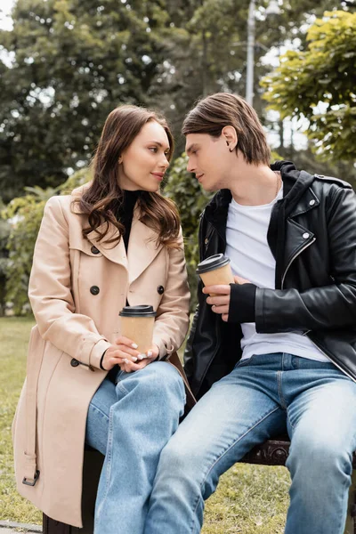 Jeune et élégant couple tenant des tasses en papier avec boisson à emporter tout en étant assis sur le banc dans le parc — Photo de stock