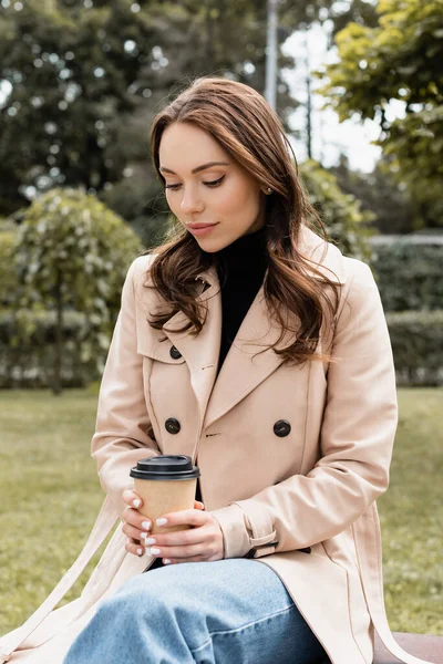 Jeune femme en trench coat beige assise sur banc avec tasse en papier — Photo de stock