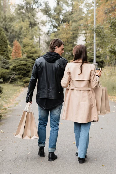 Vue arrière de la femme en trench coat tenant tasse en papier près du petit ami avec des sacs à provisions tout en marchant dans le parc — Photo de stock