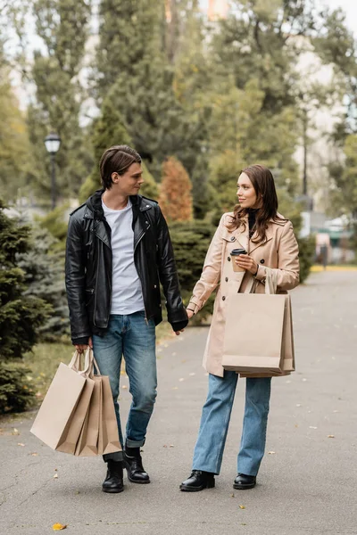 Visão comprimento total da mulher segurando copo de papel e mão do namorado com sacos de compras no parque — Fotografia de Stock