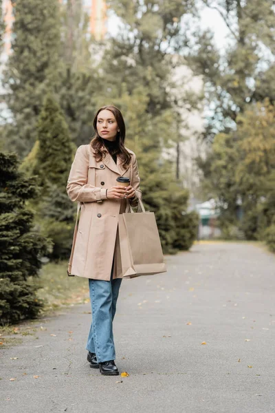 Вид в полный рост молодой женщины в трикотажном пальто, держащей бумажный стаканчик и сумки для покупок в парке — стоковое фото