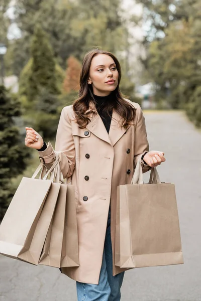 Bonita mujer joven en gabardina sosteniendo bolsas de compras en el parque otoñal - foto de stock