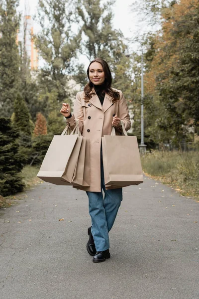 Longitud completa de la joven feliz en gabardina sosteniendo bolsas de compras en el parque otoñal - foto de stock