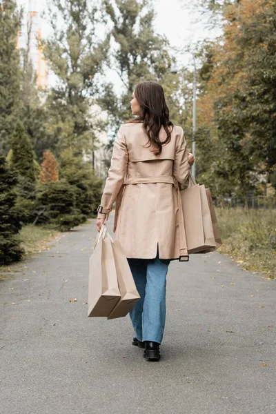 Piena lunghezza di giovane donna in trench che tiene borse della spesa e cammina nel parco autunnale — Foto stock