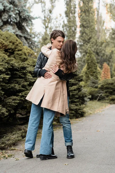Comprimento total de jovem dançando com namorada morena em casaco de trincheira no parque outonal — Fotografia de Stock