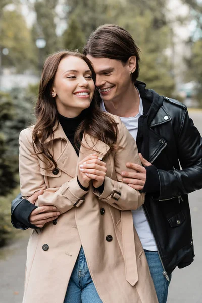 Joyful young man hugging happy girlfriend in trench coat outdoors — Stock Photo