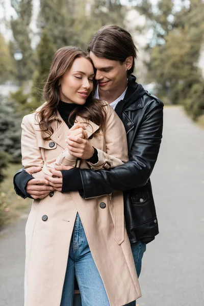 Щасливий молодий чоловік обіймає задоволену дівчину в траншеї пальто на відкритому повітрі — стокове фото