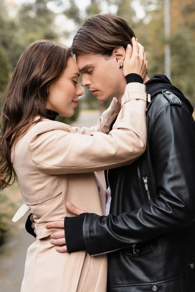 Vista lateral de mujer complacida en gabardina beige abrazando novio en chaqueta negra - foto de stock