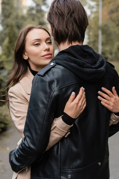 Mujer joven y bonita en abrigo beige abrazando novio en chaqueta negra - foto de stock