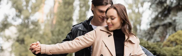 Hombre alegre cogido de la mano con la novia complacida en gabardina beige en el parque, pancarta - foto de stock