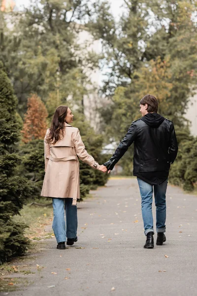 Longitud completa de pareja joven y elegante en trajes otoñales tomados de la mano mientras camina en el parque - foto de stock