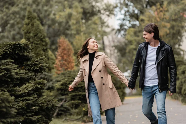 Pareja alegre y elegante en trajes otoñales tomados de la mano mientras camina en el parque - foto de stock