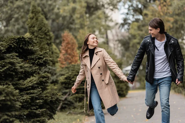 Alegre y elegante pareja en trajes otoñales tomados de la mano mientras camina en el parque - foto de stock