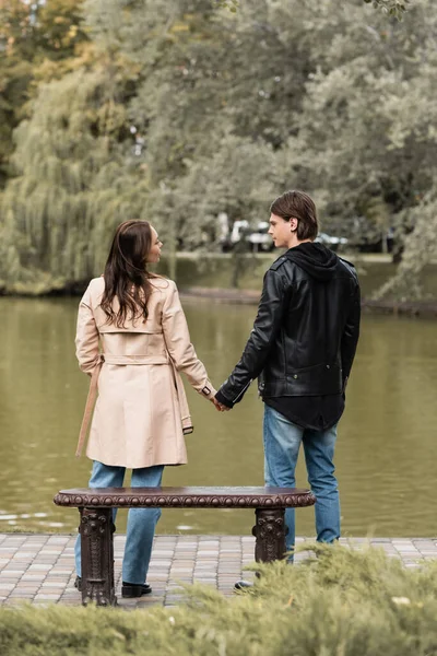 Back view of young couple in stylish outfits holding hands while standing near lake in autumnal park — Stock Photo