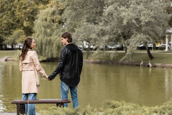 Pareja joven en trajes otoñales tomados de la mano mientras están cerca del lago en el parque - foto de stock