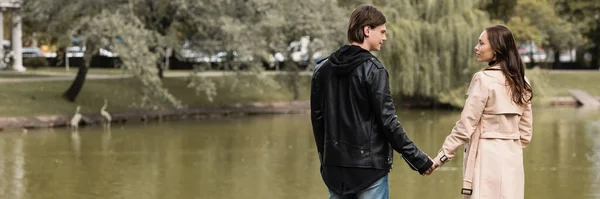 Молода пара в автентичному вбранні тримає руки, стоячи біля озера в парку, банер — стокове фото