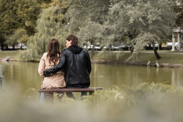 Vista posteriore del giovane in abito autunnale abbracciare fidanzata mentre seduto sulla panchina vicino al lago nel parco — Foto stock