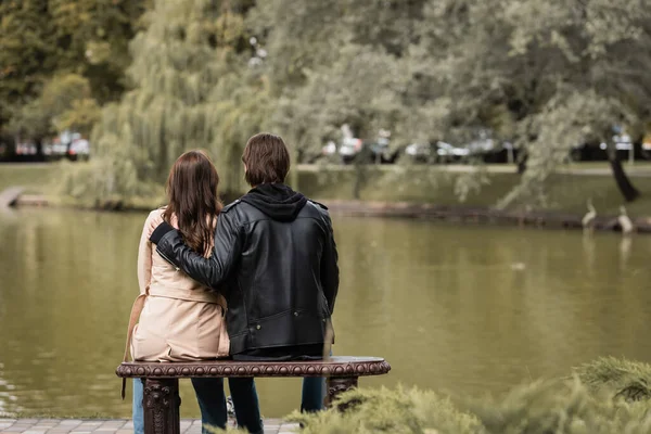 Back view of young man in black jacket hugging girlfriend while sitting on bench near lake in park — Stock Photo