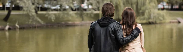 Vista trasera de hombre joven en chaqueta negra abrazando novia cerca del lago en el parque, pancarta - foto de stock