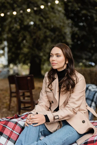 Dreamy young woman in beige trench coat sitting on blanket during picnic in park — Stock Photo