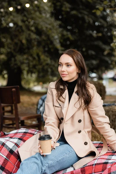 Joyeuse jeune femme en trench coat beige assise sur une couverture et tenant une tasse en papier — Photo de stock