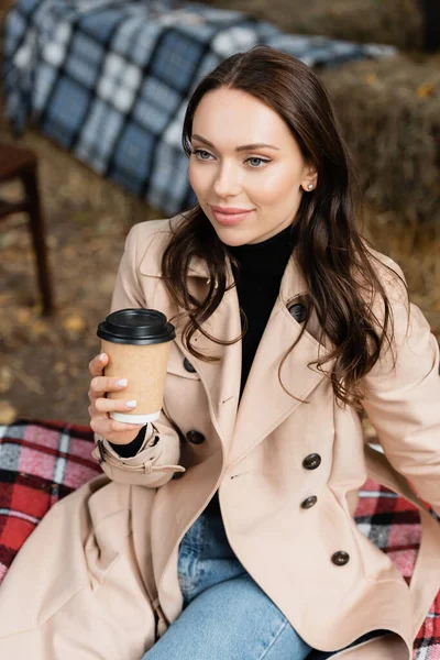 Pleased young woman in beige trench coat sitting on blanket and holding paper cup — Stock Photo