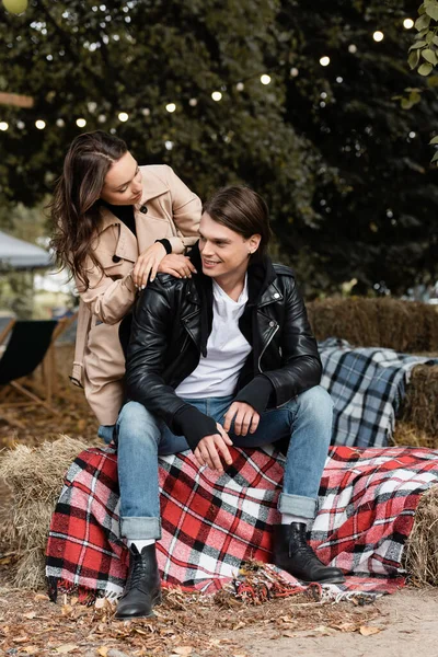 Jeune femme en trench coat regardant l'homme joyeux assis sur la couverture dans le parc — Photo de stock