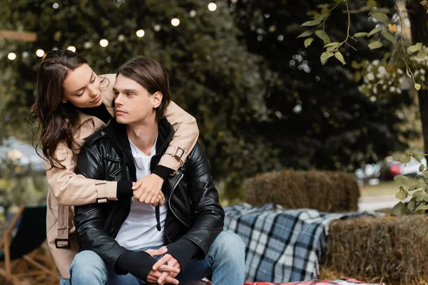 Young woman in trench coat hugging stylish boyfriend sitting with clenched hands in park — Stock Photo