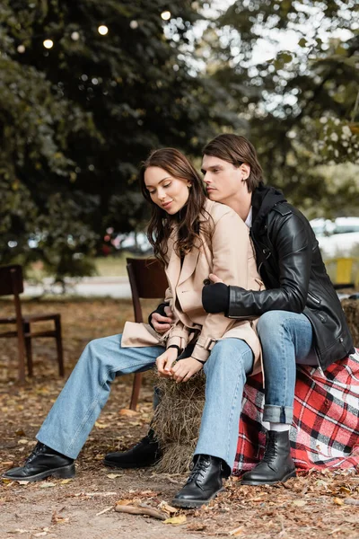 Stylish man in black jacket hugging young girlfriend in trench coat sitting in park — Stock Photo