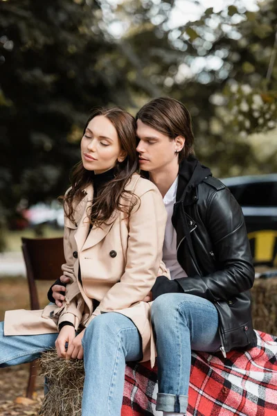 Stylish man in black jacket hugging young girlfriend with closed eyes sitting in park — Stock Photo