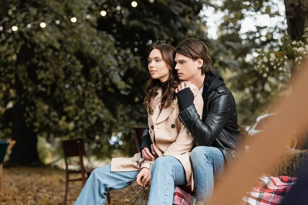 Hombre con estilo en chaqueta abrazando joven novia en abrigo beige sentado en el parque - foto de stock