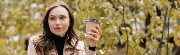 Femme rêveuse en trench coat beige tenant tasse en papier avec café pour aller dans le parc automnal, bannière — Photo de stock