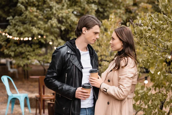 Vista lateral de la mujer joven y el hombre en trajes elegantes que sostienen el café para ir y mirarse en el parque - foto de stock