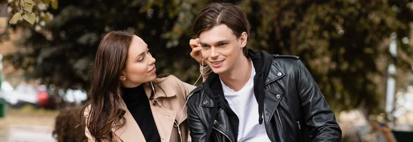 Smiling young woman adjusting wired earphone in ear of happy boyfriend, banner — Stock Photo