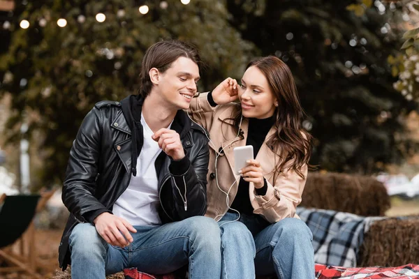 Alegre joven mujer sosteniendo teléfono inteligente mientras escucha música y compartir auriculares con cable con el novio - foto de stock