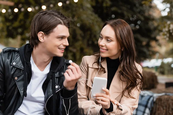 Alegre joven mujer sosteniendo teléfono inteligente mientras escucha música y compartir auriculares con cable con novio feliz - foto de stock