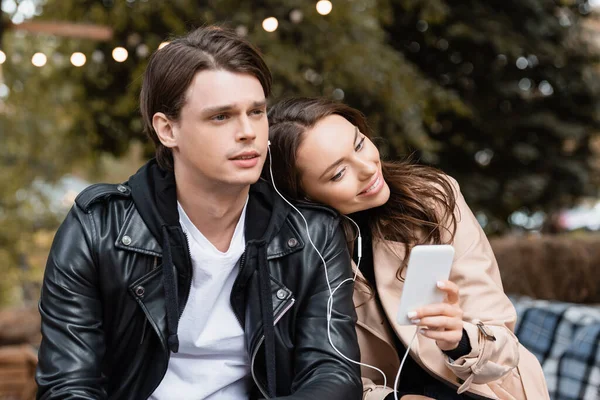 Cheerful woman holding smartphone while sharing wired earphones with boyfriend and listening music together — Stock Photo
