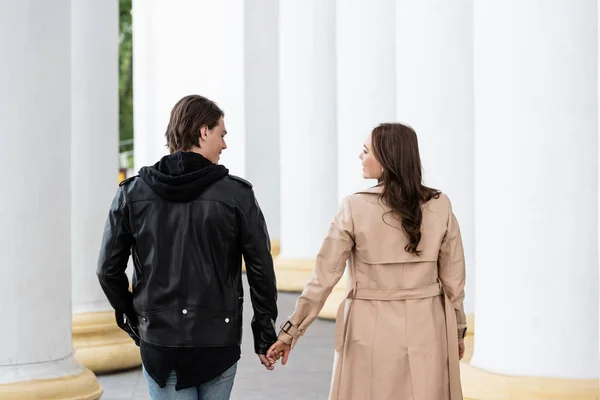 Feliz joven hombre y mujer tomados de la mano mientras camina cerca de columnas blancas - foto de stock