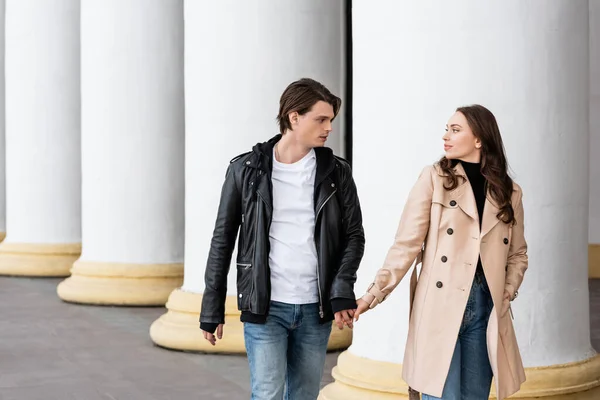 Young man and woman in trench coat holding hands while walking near white columns — Stock Photo