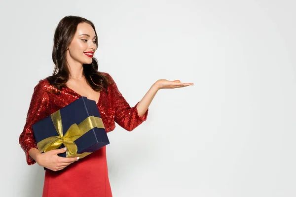 Mulher morena sorridente em vestido vermelho segurando presente e apontando com a mão isolada no cinza — Fotografia de Stock