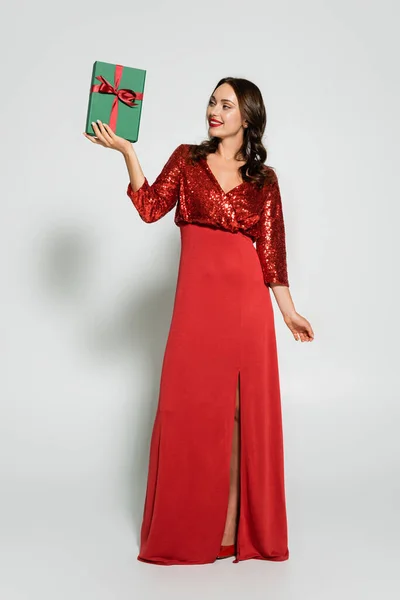 Comprimento total da mulher elegante em vestido vermelho olhando para a caixa de presente no fundo cinza — Fotografia de Stock