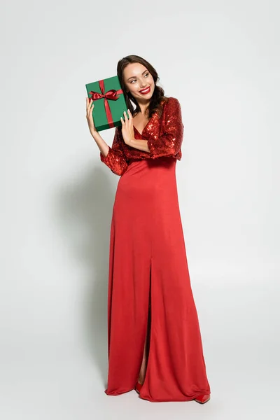 Longitud completa de la mujer bonita en vestido rojo celebración de regalo con lazo sobre fondo gris - foto de stock