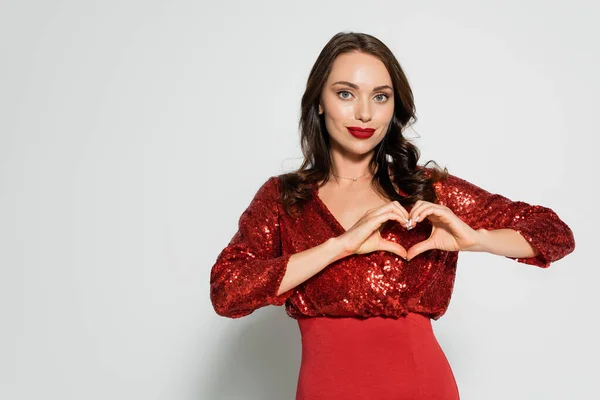 Stylish brunette woman in red dress showing heart gesture on grey background — Stock Photo